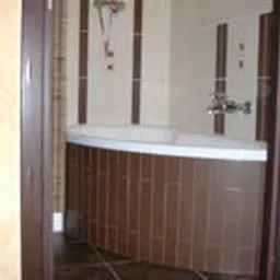 Narożna wanna obudowana brązowymi płytkami, widoczna zza framugi drzwi w wykończonej łazience z beżowo-brązowymi akcentami.